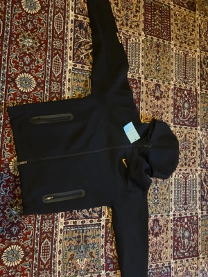 Svart Nike hoodie med dragkedja - Svart hoodie från Nike med hel dragkedja framtill och två vertikala fickor med dragkedja. Klassisk huva och gul Nike-logga på bröstet. Perfekt för en sportig och avslappnad stil. Ingen papper ine i den 