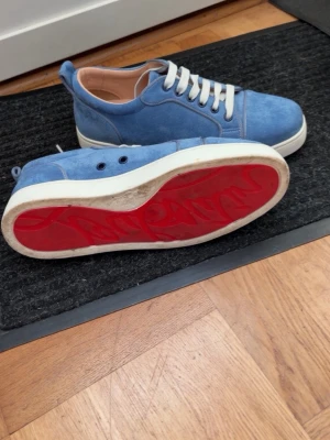 Blåa sneakers från Christian Louboutin - Snygga blåa sneakers i mocka från Christian Louboutin med klassisk röd sula och vita skosnören. Låg modell med rund tå och diskreta detaljer. Perfekta för dig som vill sticka ut med exklusiva skor och unik design.