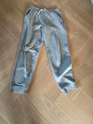 Grå mjukisbyxor från Polo Ralph Lauren - Snygga grå mjukisbyxor från Polo Ralph Lauren med dragsko i midjan och två sidofickor. Klassisk broderad logga i mörkblått på vänster sida. Tillverkade i mjukt bomullsmaterial och har ribbade muddar vid bensluten för en skön passform