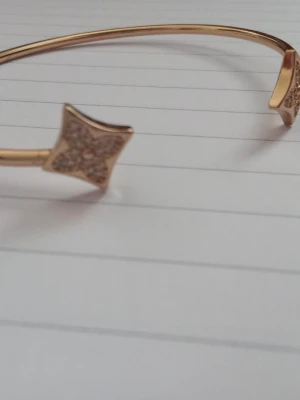 Guldfärgat armband med stjärndetaljer - Snyggt guldfärgat armband i metall med två stjärnformade detaljer i varje ände. Stjärnorna är dekorerade med små glittrande stenar som ger extra bling. Armbandet har inte använts och har inga märken från användning. Jag vet inte vad det är för sorts metall den är gjord av.