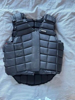 Svart skyddsväst från Jacson (för barn) - Säljer en svart skyddsväst från Jacson med vadderade paneler och justerbara kardborreband i sidorna. Västen har en sportig look och är tillverkad i slitstarkt syntetmaterial. Perfekt för dig som vill ha extra skydd vid ridning eller annan aktivitet.