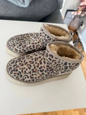 Leopardmönstrade UGG boots - Säljer ett par snygga UGG boots med leopardmönster och beige botten. Bootsens insida är fodrad med mjuk, beige fuskpäls som håller fötterna varma. Yttermaterialet är mocka och sulan är i gummi. Perfekta för dig som vill sticka ut med ett djurmönster.