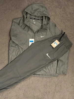 Nike miller - Snyggt grått träningsset från Nike med matchande jacka och byxor. Jackan har huva och dragkedja framtill, medan byxorna har resår i midjan och dragsko. Båda plaggen har Nike-logga i vitt och är tillverkade i ett lätt, sportigt material.