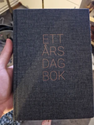 Ett års dagbok i tygklädd design - Snygg dagbok med titeln 'Ett års dagbok' i kopparfärgad text på ett mörkgrått tygklätt omslag. Perfekt för att skriva ner tankar, mål eller minnen under ett helt år. Stilren och enkel design som passar på skrivbordet eller i väskan.