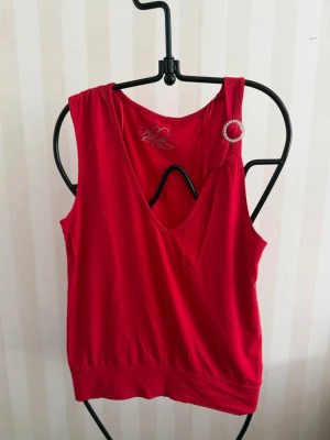 Röd linnetopp med dekorativ brosch - Snygg röd linnetopp från systers  med djup v-ringning och en dekorativ broschdetalj på axeln. Toppen är ärmlös och har bredare axelband. Perfekt för dig som vill sticka ut lite extra. Materialet känns mjukt och följsamt.