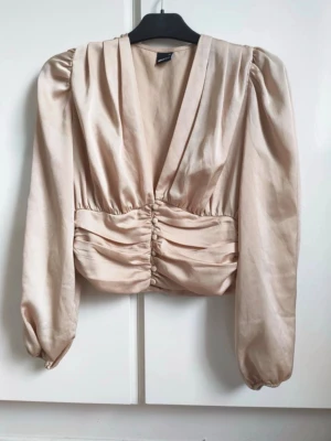 Champagnefärgad satinblus med v-ringning - Snygg champagnefärgad blus från NA-KD i glansigt satinmaterial. Djup v-ringning, puffiga långärmar och markerad midja med dekorativa knappar framtill. Perfekt för dig som vill ha en elegant och trendig look.