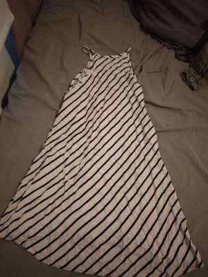 Vit och svart randig långklänning - Långklänning med tunna axelband i vitt tyg med svarta diagonala ränder. Klänningen har en lös passform och är perfekt för varma dagar. Materialet känns lätt och luftigt, troligen bomull eller polyester.