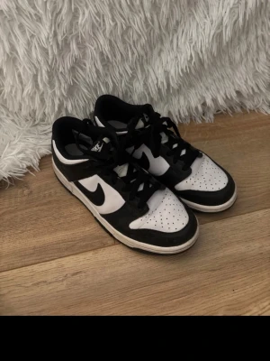 Nike Dunk Low svart/vit sneakers - Nike Dunk Low sneakers i svart och vitt med klassisk Swoosh-logga på sidan. Skorna har lågt skaft, snörning och ovandel i läder. Perfekt för dig som gillar streetwear och vill ha en tidlös look.