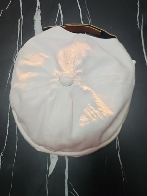 Vit streetwear keps med brodyr - Säljer en vit keps i streetwear-stil med rundad form och uppvikt kant. På baksidan finns en orange patch med svart broderad text 'without guts talent is waste'. Kepsen är tillverkad i tyg och har en knapp på toppen.