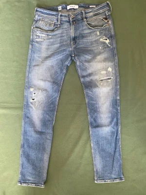 Blå slitna jeans från Replay - Säljer ett par ljusblå jeans från Replay med slitningar och hål på både lår och knä. Jeansen har fem fickor, normal midja och raka ben. Perfekta för en avslappnad och trendig look.