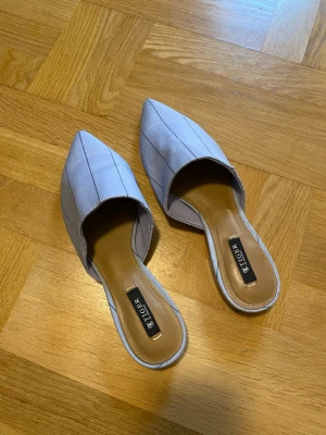 Blå slip-in sandaler från Tiger of Sweden - Säljer ett par stilrena slip-in sandaler från Tiger of Sweden i ljusblått tyg med tunna vertikala ränder. Skorna har spetsig tå och öppen häl, samt en mjuk innersula i beige. Perfekta för en trendig och avslappnad look!
