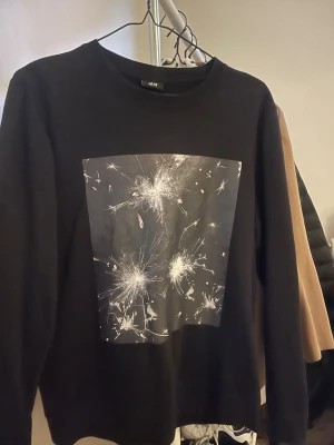 Svart långärmad tröja med tryck - Svart långärmad tröja med ett stort grafiskt tryck framtill som liknar fyrverkerier eller stjärnexplosioner i vitt och grått. Tröjan har rund halsringning och är i mjukt material, perfekt för en cool och avslappnad stil. Säljs för 30 kr 