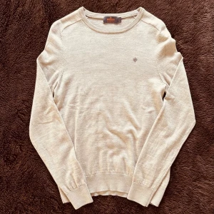 Morris beige tröja - Stilren beige tröja från Morris med rund halsringning och diskret broderad logga på bröstet. Tillverkad i 100% merinoull och har ribbade muddar vid ärmslut och nederkant. Perfekt för kallare vårdagar/sommarkvällar och enkel att matcha. Nypris cirka 1600kr!