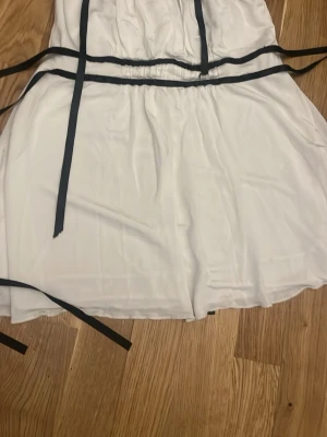 Vit top med svarta band - Säljer en vit top med svarta knytband i midjan och längs sidorna. Toppen är ärmlös och har en loose passform med flowy tyg, troligtvis i polyester. Enkel men stilren design som sticker ut med sina kontrasterande detaljer.