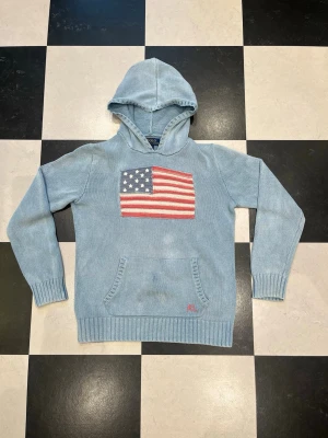 Ljusblå hoodie från Polo Ralph Lauren - Ljusblå hoodie från Polo Ralph Lauren med amerikansk flagga broderad på bröstet. Klassisk känguruficka framtill och ribbade muddar vid ärmar och nederkant. Tillverkad i mjukt stickat material och har en avslappnad passform.