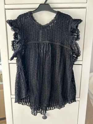 Svart spetsblus med volangärmar - Snygg svart blus i spets med volangdetaljer vid ärmarna och rund halsringning. Blusen har knytning i nacken och är lätt transparent med ett luftigt fall. Perfekt för dig som gillar en romantisk och trendig stil. Passar både XXS och XS💗