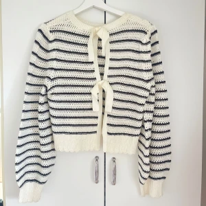 Vit stickad kofta med svarta ränder - Säljer en söt vit stickad kofta med svarta horisontella ränder och två knytband framtill. Koftan har ballongärmar och ribbade muddar vid ärmslut och nederkant. 