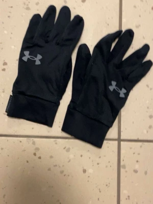Svarta träningshandskar Under Armour - Svarta, tunna träningshandskar från Under Armour med logga i vitt på ovansidan. Perfekta för löpning eller utomhusträning när det är lite kyligt. Sitter tight runt handleden och har en sportig look.