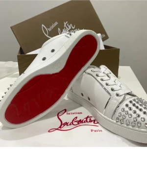 Vita Christian Louboutin sneakers med nitar - Snygga vita sneakers från Christian Louboutin med ikonisk röd sula och silverfärgade nitar på tå och häl. Skorna har en klassisk låg siluett, vita skosnören och är tillverkade i slätt läder. Perfekta för dig som vill sticka ut med en edgy och lyxig stil.