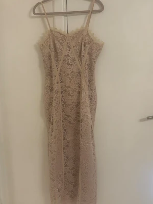 Beige spetsklänning med smala axelband - Säljer en elegant beige spetsklänning med smala axelband och figurnära passform. Klänningen har vackra spetsdetaljer och är vadlång, perfekt för dig som gillar en feminin och stilren look. Passar till sommarens alla tillfällen!