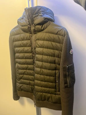Olivgrön pufferjacka från Moncler - Snygg olivgrön pufferjacka från Moncler med stickade ärmar och detaljer. Jackan har huva, dragkedja framtill och fickor med tryckknappar. Ärmen har en extra ficka med dragkedja och Moncler-logga. Perfekt för kalla dagar och riktigt trendig look. Stl L passar m