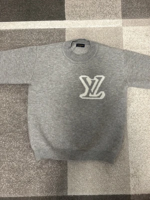 Louis Vuitton tröja Grå - Snygg grå stickad tröja från Louis Vuitton med stor vit LV-logga på bröstet. Tröjan har rund halsringning, ribbade muddar och är tillverkad i ett mjukt material som känns lyxigt. Perfekt för dig som gillar exklusiva streetwear-plagg.