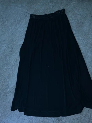 Svart långkjol med resårmidja - En svart långkjol med bred resår i midjan som har små dekorstenar för extra detalj. Kjolen är lätt och något genomskinlig, vilket ger en luftig känsla. Perfekt att styla med en crop top eller stickad tröja.