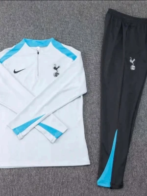 Tottenham träningsset från Nike - Snyggt Tottenham Hotspur träningsset från Nike med långärmad vit tröja och svarta byxor. Tröjan har blå detaljer och halv dragkedja, byxorna har blå inslag vid benslut. Båda plaggen har klubbens logga tryckt på framsidan.