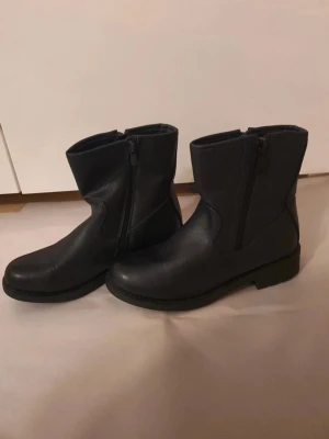 Svarta boots med dragkedja - Snygga svarta boots i skinnimitation med grov sula och dragkedja på sidan. Klassisk modell med rund tå och mellanhögt skaft, perfekta till höst- och vinteroutfits. Passar dig som gillar en stilren och enkel look.