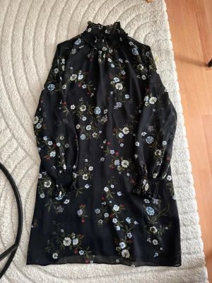 Svart blommig klänning med spets - Svart kortklänning med blommigt mönster i vitt, blått och grönt. Klänningen har långärmade ärmar med smock vid handlederna och en hög krage med spetsdetaljer. Perfekt för dig som gillar romantiska och trendiga plagg.