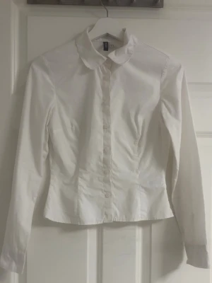 Vit figursydd skjorta från H&M - Säljer en klassisk vit skjorta från H&M med figursydd passform och krage. Skjortan har långa ärmar och knappar hela vägen framtill. Perfekt för dig som vill ha en stilren och enkel look. Pris kan diskuteras vid snabbt köp och ett litet märke vid bakre axeln men syns inte när den är på. Den är str xs/xxs. Endast andvänd under min praktik i en väcka.