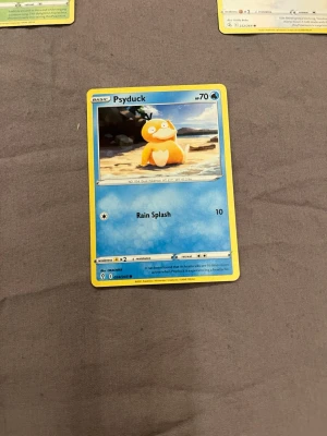 Pokémonkort Psyduck - Säljer ett original Pokémonkort med Psyduck-motiv. Kortet har blå och gul ram, och visar Psyduck på en strand. Texten är på engelska och kortet har attacken Rain Splash. Perfekt för samlare eller Pokémonfans.