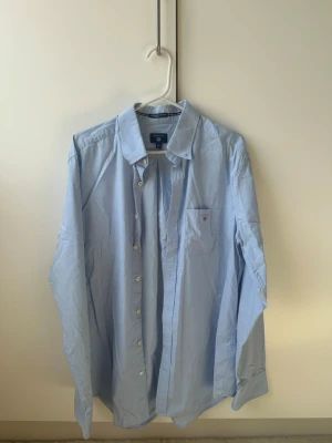 Ljusblå skjorta från Gant - Klassisk ljusblå skjorta från Gant med lång ärm och knappar framtill. Skjortan har en bröstficka med liten logga och är tillverkad i bomull. Perfekt till jeans eller chinos för en clean look.