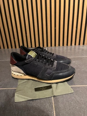 Valentino  Garavani Rockrunners - Snygga Valentino Garavani Rockrunner  i mörkblått och beige med vinröda detaljer på hälen. Ovandelen är i mesh och mocka, med skinnpaneler och klassisk snörning. . Perfekt för dig som gillar exklusiva sneakers med unik design. Skick 7/10 då ena valentino lappen är borta. Kommer även med dustbag, om ni funderar över något så är det bara att fråga. 