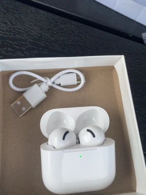 Trådlösa vita hörlurar med laddningsetui - Säljer ett par trådlösa vita hörlurar med stilren design och laddningsetui. Hörlurarna har en slät, rundad form och levereras med en USB-laddningskabel. Perfekta för musik, poddar och samtal på språng.