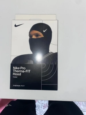 Nike Pro Therma-FIT Hood - Nike Pro Therma-FIT Hood är en svart huva designad för att hålla dig varm under träning eller match. Tillverkad i ett stretchigt och isolerande material som andas, perfekt för fotboll eller andra sporter. Snygg och funktionell design med Nike-logga.