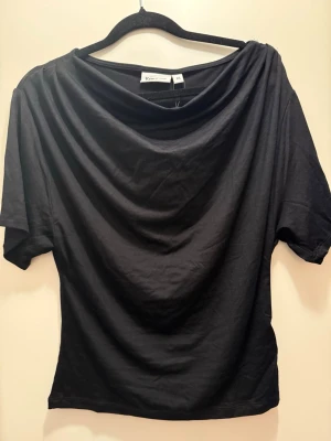 Svart draperad topp från Kappahl - Svart topp från Kappahl med draperad halsringning och korta ärmar. Tillverkad i mjuk bomull som känns skön mot huden. Enkel och stilren design som passar till många olika looks.