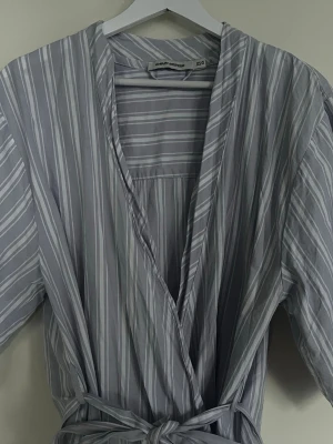 Randig morgonrock från Carin Wester  - Ljusgrå morgonrock med vita och ljusblåa ränder från Carin Wester. Modellen har knytskärp i midjan och lös passform, perfekt för att chilla hemma. Tunn och mjuk känsla i materialet.