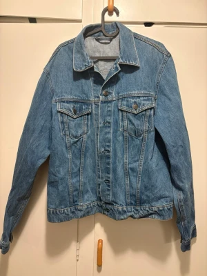 Klassisk blå jeansjacka - Säljer en klassisk blå jeansjacka i tidlös modell med två bröstfickor med lock och knapp, samt knappar framtill. Jackan har en rak passform, skjortkrage och justerbara knappar vid ärmslut och midja. Perfekt att slänga över en hoodie eller t-shirt.