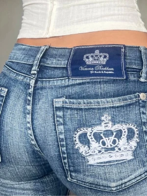Victoria Beckham blå bootcut jeans - Säljer ett par blå bootcut jeans från Victoria Beckham x Rock & Republic. Jeansen har en snygg tvätt, låg midja och vita broderade kronor på bakfickorna. Klassisk femficksmodell med silverfärgade detaljer och slitningar för en cool look.