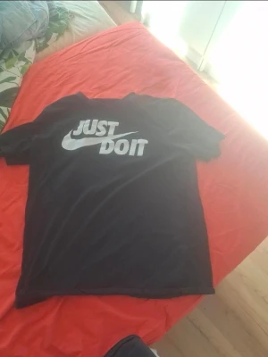 Svart Nike Just Do It t-shirt - Säljer en svart t-shirt från Nike med det klassiska 'Just Do It'-trycket i vitt på bröstet. T-shirten har korta ärmar och rund hals. Perfekt för dig som gillar sportig och enkel stil.