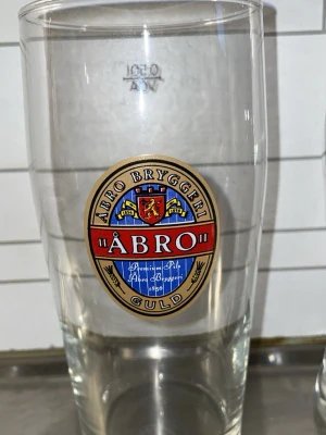 Åbro Bryggeri ölglas - Säljer ett klassiskt ölglas från Åbro Bryggeri med färgglad logga i rött, blått och guld. Glaset är genomskinligt och har en rund form med tryckt emblem på framsidan. Perfekt för dig som gillar samlarprylar eller vill ha snygga glas till festen.
