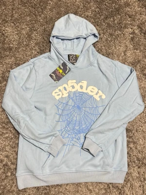 Ljusblå hoodie från Sp5der - Snygg ljusblå hoodie från Sp5der med vit logga i 3D-tryck och ett stort blått spindelnätsmönster på framsidan. Hoodien har huva och magficka, samt ribbade muddar vid ärmslut och nederkant. Perfekt för dig som gillar streetwear och vill sticka ut.