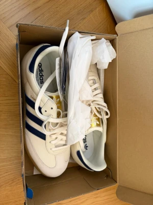Adidas Samba OG vita och marinblå - Säljer ett par Adidas Samba OG sneakers i vitt läder med klassiska marinblå ränder och gulddetaljer på plösen. 