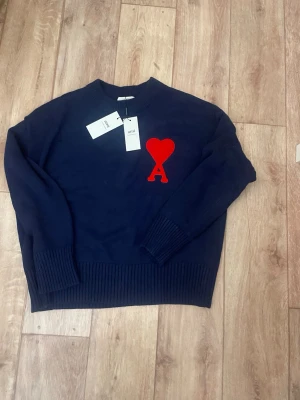 Mörkblå stickad tröja från Ami - Snygg mörkblå stickad tröja från Ami Alexandre Mattiussi med röd hjärtlogga och bokstaven A på bröstet. Tröjan har rund hals, ribbade muddar och nederkant. Perfekt för dig som gillar stilrena detaljer och trendiga plagg.