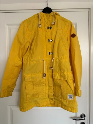 Gul vindjacka från Dobber - Gul vindjacka från Dobber med huva och vita dragskor. Jackan har träknappar, flera fickor framtill och ett brunt patchmärke på ärmen. Storlek XS, välanvänd så därav några defekter som inte påverkar helhetsintrycket särskilt mycket men bra att veta (se bilder). Tillverkad i slitstarkt material🤗