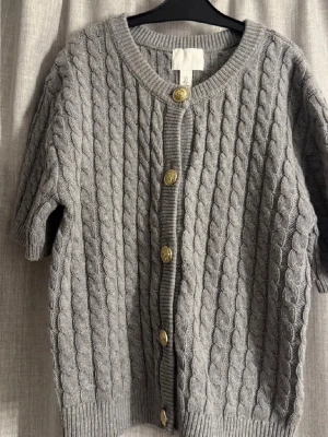 Grå kabelstickad kofta H&M - Kortärmad grå kabelstickad kofta från H&M med rund halsringning och dekorativa guldfärgade knappar framtill. Koftan har ribbade muddar och ett klassiskt mönster som ger en trendig look. Perfekt att styla över en topp eller klänning.