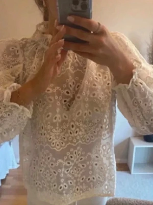 Vit broderad blus med volang - Säljer en superfin vit blus från Gina Tricot med broderade detaljer och volang vid halsen. Blusen har lång ärm och är gjord i ett tunt, luftigt material med hålmönster. Perfekt för dig som gillar romantisk och trendig stil.