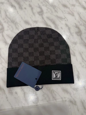 En ljus svart mössa - En ljus svart stickad mössa med rutigt mönster och uppvikt kant, med en liten logotyp framtill och etikett kvar.