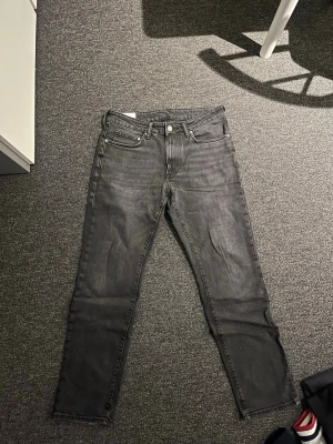 Grå raka jeans - Säljer ett par grå jeans med rak passform och klassisk femficksdesign. Jeansen har normal midja och stängs med knapp och dragkedja. Materialet är denim i bomull och de har en stilren, enkel look som passar till det mesta.